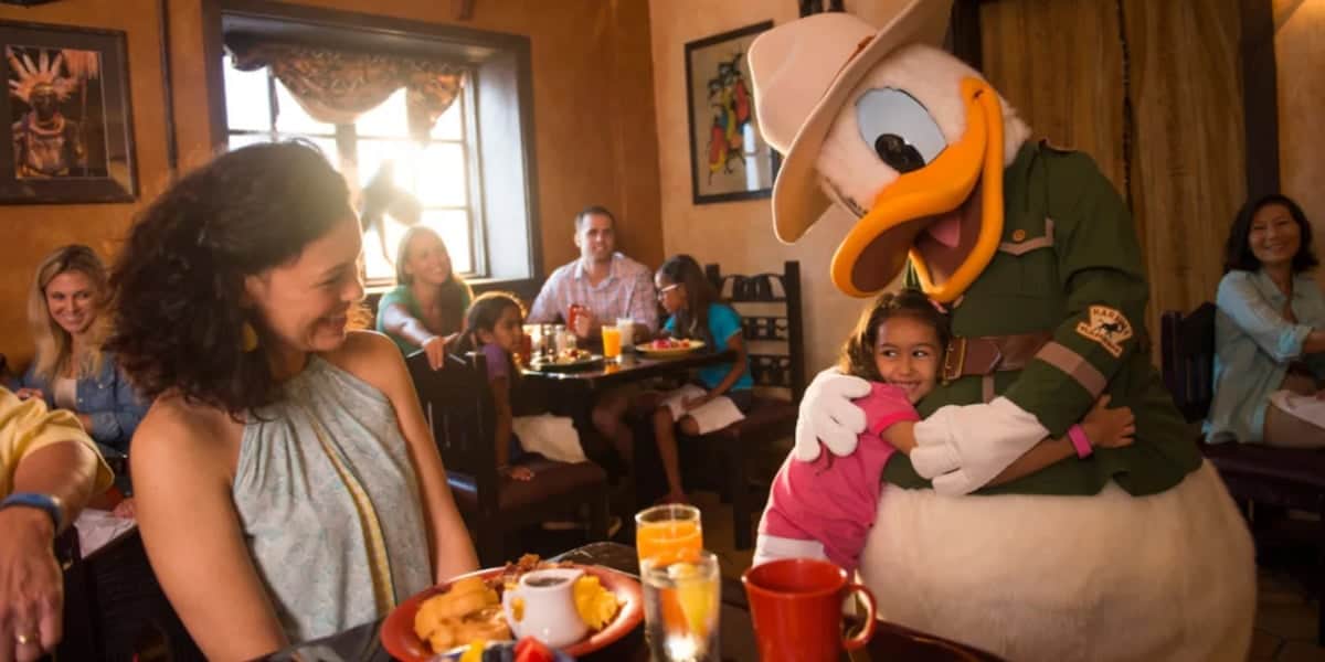 A young girl hugs a Disney character dressed as a park ranger at a restaurant. Smiling adults and children sit at tables with plates of food, and another child watches the interaction. The atmosphere is lively and warm, with decorations and sunlight coming through the window.