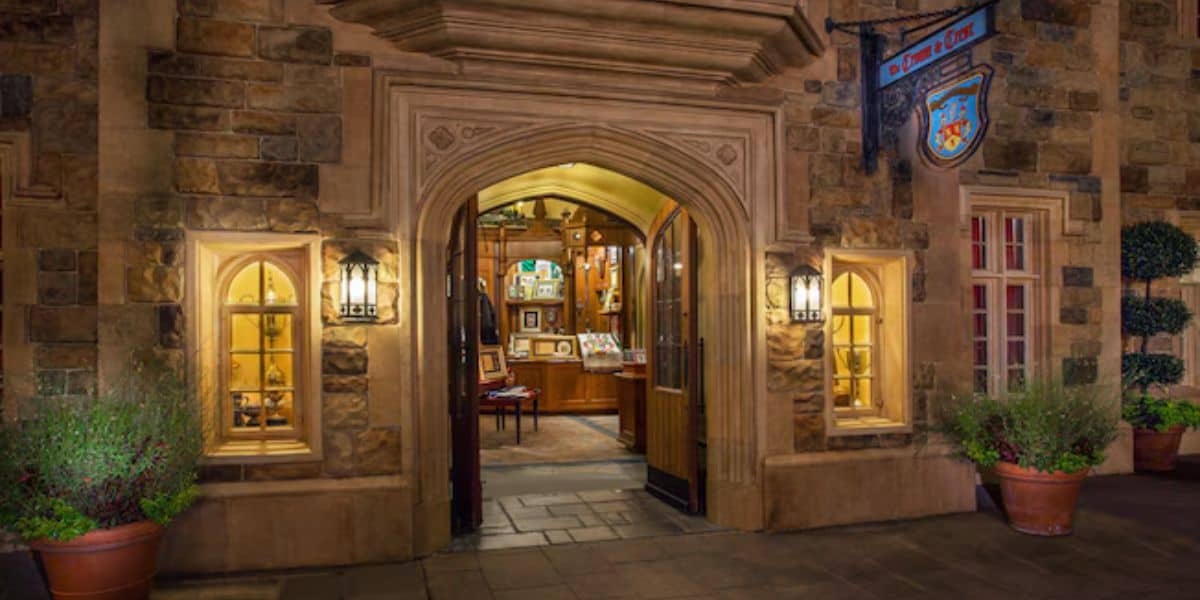 A charming, warmly-lit store entrance with arched stone doorways and wooden double doors, flanked by windows and potted plants. Inside, shelves and walls are adorned with various framed pictures and collectibles, creating an inviting and cozy atmosphere.
