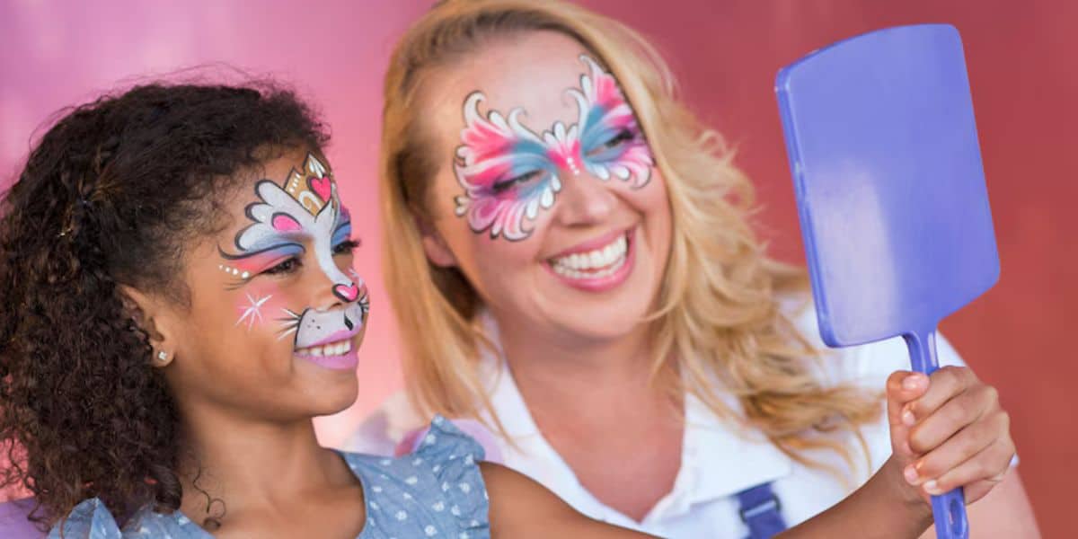 Two women with colorful face paint smile. The girl, painted as a bunny with flowers, holds up a purple hand mirror. The older woman, painted with elaborate butterfly designs, looks at her reflection, creating a joyful moment against a pink background that almost feels like magic under the Disney dress code.