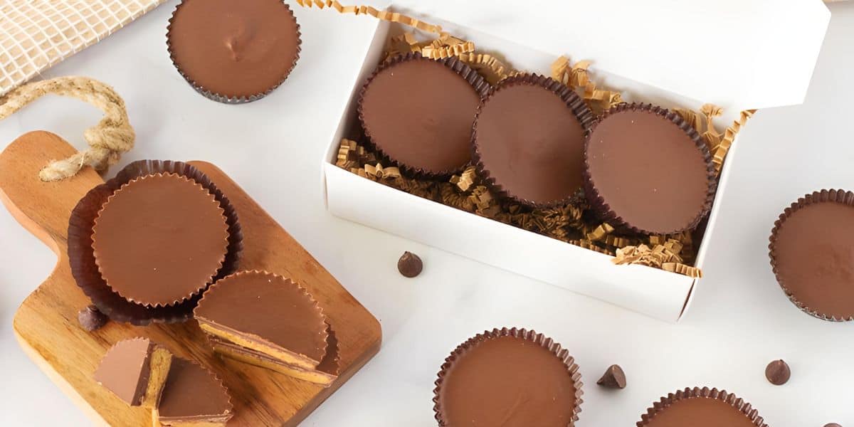 A selection of homemade chocolate cups are displayed on a table. Some are in a white box filled with decorative paper shreds, while others are arranged on a small wooden cutting board and plates. Chocolate chips are scattered around as decoration.