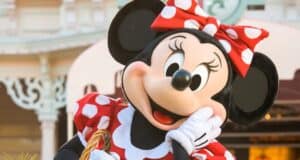 Minnie Mouse, dressed in her classic red and white polka-dot dress with a matching bow, poses cheerfully with one hand on her cheek. She stands in front of a whimsical building, likely within a theme park setting, exuding joy and playfulness.