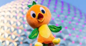 A person holds up a plush toy of an animated orange bird with green leaves as wings and on its head. The background, reminiscent of Disney World, features a futuristic geodesic dome structure with pink lights.