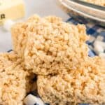 A stack of rice crispy treats sits on a blue and white checkered cloth. Scattered mini marshmallows surround the treats, and a stick of butter is visible in the background. A glass dish with more treats is partially shown on the right side of the image.