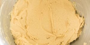 A close-up view of a mixing bowl filled with creamy, light brown cookie dough. The dough looks smooth and evenly mixed, ready for baking. The bowl appears to be made of metal.