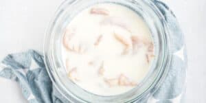 A glass bowl filled with raw chicken pieces marinating in a white liquid, likely buttermilk, sits on a kitchen counter. The bowl is placed on a blue and white patterned cloth. The image is taken from above, showcasing the ingredients.
