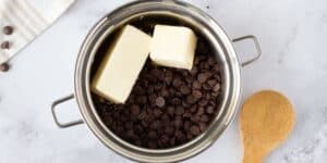 A metal saucepan with two sticks of butter and a pile of chocolate chips inside sits on a marble countertop. A wooden spoon lies nearby, and a white and striped cloth is partially visible in the upper left corner.