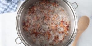 A pot filled with small pieces of bacon frying in bubbling oil, setting the stage for a cheesy potato soup that rivals Le Cellier’s iconic dish. A wooden spoon rests beside the pot on a white countertop, with a blue and white checkered cloth in the background.