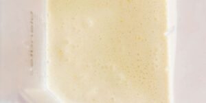 A close-up view of a blender with pale yellow, frothy liquid inside, likely a smoothie or milkshake, against a light background. Only part of the blender's side with measurement markings is visible, highlighting the creamy texture of the contents.