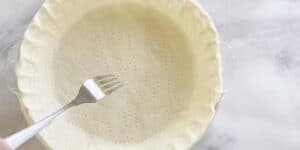 A close-up of an uncooked pie crust in a glass pie dish set on a marble countertop. The crust is docked with small holes made by a fork, which is held by a hand resting on the edge of the dish.