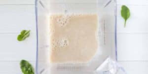 A top-down view of a blender containing a creamy white liquid, possibly a smoothie. The blender is positioned on a white surface with a few spinach leaves scattered around it.