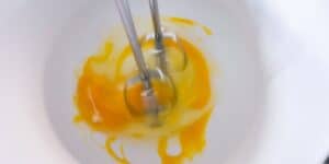 A close-up image shows an electric mixer blending egg yolks in a white mixing bowl. The mixer blades are in motion, creating a swirl of yellow and white as the ingredients mix together.