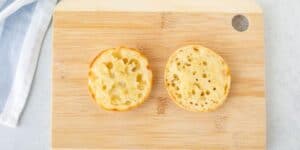 Two halves of an English muffin with a light golden crust and characteristic nooks and crannies are placed face up on a wooden cutting board. A blue and white cloth is partially visible on the left side of the board.