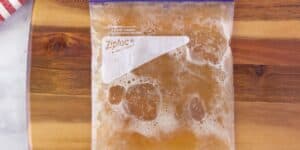 A transparent Ziploc bag filled with a tan-colored liquid and some bubbles is placed on a wooden surface. The bag has a white label, and part of a red-and-white striped cloth is visible in the top left corner of the image.