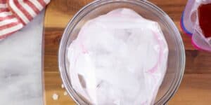 A clear plastic bag filled with ice cubes sits in a transparent bowl on a wooden cutting board. There’s a red and white striped cloth at the edge of the board on the left, and a cut-off view of another bag with red contents to the right.