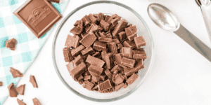 A glass bowl filled with chopped chocolate pieces sits on a white surface. To the left, a checkered teal and white cloth cradles a bar of chocolate. To the right, a metal ice cream scoop is visible, hinting at dessert preparation for a Frozen Hot Chocolate Recipe as delightful as Olaf!