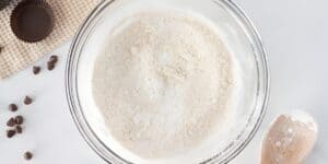 A clear glass mixing bowl filled with flour mixture is placed on a white countertop. To the left are chocolate chips, a chocolate cup, and a cloth. A wooden spoon with flour rests on the right side near the bowl.