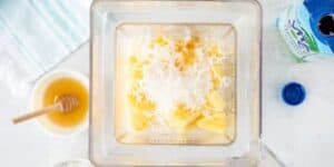 A top-down view of a blender filled with pineapple chunks, shredded coconut, and a light liquid. A small bowl with honey and a honey dipper is on the left, along with a partially opened can of coconut water and a striped towel.