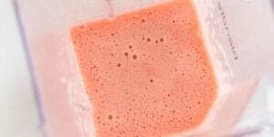 A close-up view of a blender container filled with freshly blended pink smoothie, with small bubbles visible on the surface. The blender walls are slightly frosted, and the smoothie appears creamy and smooth.