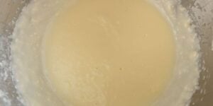 A close-up view of a smooth, yellow mixture in a glass bowl, likely a batter or a thick custard. The mixture appears creamy and well-blended, with a few bubbles visible on the surface. The bowl is partially seen, and the mixture reaches halfway up the sides.