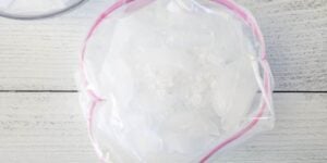 A top-down view of a pink and white bowl covered with plastic wrap containing translucent, ice-like contents. The bowl is placed on a white wooden surface, and part of another transparent lid is visible in the upper left corner of the image.