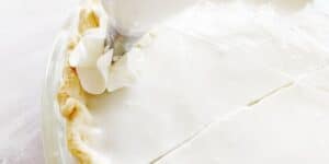 A close-up shows a partially sliced pie with a creamy, smooth white top. A piping bag is adding decorative whipped cream swirls along the edge of the pie crust.