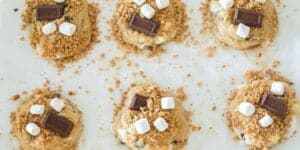 A baking sheet holding six unbaked cookies is shown. Each cookie is topped with melted chocolate pieces, marshmallows, and a sprinkling of crushed graham crackers. They are spaced evenly on the parchment paper-lined sheet.