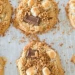 Six freshly baked s'mores cookies are arranged on a parchment-lined baking sheet. The cookies are topped with toasted marshmallows, small chocolate pieces, and crumbles of graham crackers, giving them a gooey and crunchy texture.