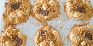 A photo of six freshly baked s'mores cookies with melted chocolate chunks, gooey marshmallows, and crumbled graham crackers on top. The cookies are placed on a parchment-lined baking sheet.