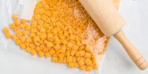 A rolling pin lying on a clear plastic bag filled with crushed orange crackers, likely cheese-flavored. The bag is partially sealed and rests on a white surface.