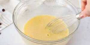 A clear glass bowl containing a yellow batter is being whisked by a hand using a metal whisk. The bowl is placed on a white countertop, and part of a white dishtowel with light stripes and a few chocolate chips are visible in the background.