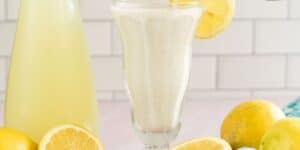 A tall glass of creamy, light yellow lemonade sits in the center, garnished with a lemon slice on the rim. Left of the glass is a matching bottle filled with more lemonade. Fresh, whole, and sliced lemons are scattered around on a light pink surface. A tiled wall is in the background.