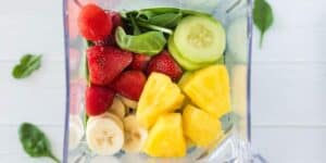 A blender pitcher filled with fresh fruits and vegetables, including sliced bananas, whole strawberries, pineapple chunks, cucumber slices, and spinach leaves, ready to be blended.