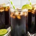 Three glass cups filled with ice and Dr Pepper, topped with lime wedges and green-striped straws, sit on a striped cloth. A can of Dr Pepper is next to the glasses, and halved limes are also nearby. The background features a white tiled wall.