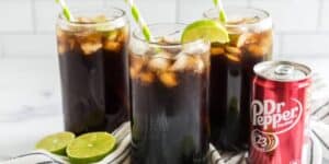 Three glass cups filled with ice and Dr Pepper, topped with lime wedges and green-striped straws, sit on a striped cloth. A can of Dr Pepper is next to the glasses, and halved limes are also nearby. The background features a white tiled wall.