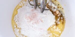 A mixing bowl containing unblended ingredients, including flour, eggs, sugar, and vanilla extract, with an electric beater partially immersed in the mixture, ready to blend the components together. The ingredients appear to be in the initial stages of preparation.