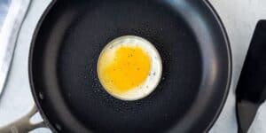 A single sunny-side-up egg frying in the center of a non-stick black skillet, surrounded by a metal ring to keep its shape. The pan handle is on the left, and a spatula handle is partially visible on the right. The background is a light-colored countertop.