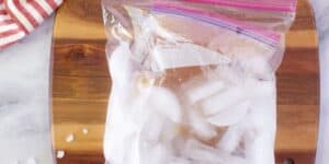 A transparent resealable plastic bag filled with ice cubes rests on a wooden cutting board. A red and white striped cloth is visible in the upper left corner. The cutting board is placed on a white marble surface with a few small ice pieces scattered around.