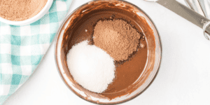 A bowl of melted chocolate with cocoa powder and sugar on top, ready to be mixed for a frozen hot chocolate recipe as delightful as Olaf! A green and white checkered cloth and a pair of measuring spoons are next to the bowl on a white surface.