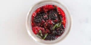 A top-down view of a glass jar containing a mixture of raspberries, blackberries, and small green leaves in a red liquid, possibly a homemade berry preserve or sauce, set against a light background.