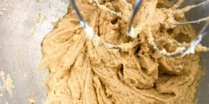 Close-up view of a mixing bowl with a peanut butter cookie dough being mixed by an electric mixer. The dough appears smooth and creamy, with the metal beater blades visible at the top of the image.