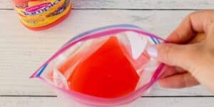A hand is holding a clear ziplock bag filled with red liquid, with a bottle of red Tampico juice partially visible in the background on a wooden surface.