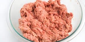 A clear glass bowl filled with a mixture of raw ground meat, likely prepared for making meatloaf or meatballs. The meat appears seasoned, showing a mix of pink and light brown colors. The bowl is placed on a white surface.