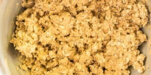 An overhead view of a mixing bowl filled with a crumbly dough mixture containing rolled oats. The oats and dough appear well-combined, giving the mixture a rough texture.