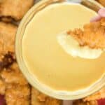 A hand holding a piece of breaded chicken nugget dipping it into a bowl of creamy yellow sauce. More chicken nuggets are scattered on a red and white checkered surface around the bowl.