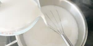 A glass measuring cup of milk is being poured into a stainless steel pot with a whisk inside. The liquid is white and foamy, indicating the beginning stages of cooking or mixing a recipe on a stovetop.