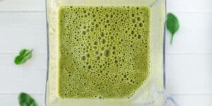 The image depicts a top-down view of a blender filled with a frothy green smoothie. There are a few spinach leaves placed around the blender on a white surface, suggesting that spinach is one of the ingredients in the smoothie.