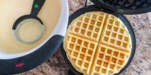 A freshly cooked waffle divided into four sections sits on a waffle maker. Beside it, a bowl filled with waffle batter is shown with a black ladle resting on top. The scene is set on a marble countertop.