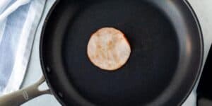 A single slice of Canadian bacon is being fried in a black nonstick pan. The pan is positioned on a light-colored surface with a folded blue and white striped cloth partially visible on the left side of the image.