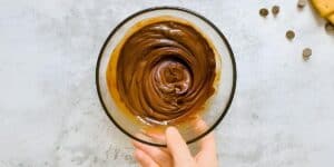 A hand holding a glass bowl filled with smooth, swirled chocolate ganache. The bowl is set on a light gray textured surface with scattered chocolate chips and a partial view of baked goods in the top right corner.
