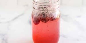 A glass jar filled with a fizzy, pink-colored liquid. The drink has small chunks of dark fruit floating on top, with bubbles visible throughout. The jar is placed on a white surface with a subtle, light background.